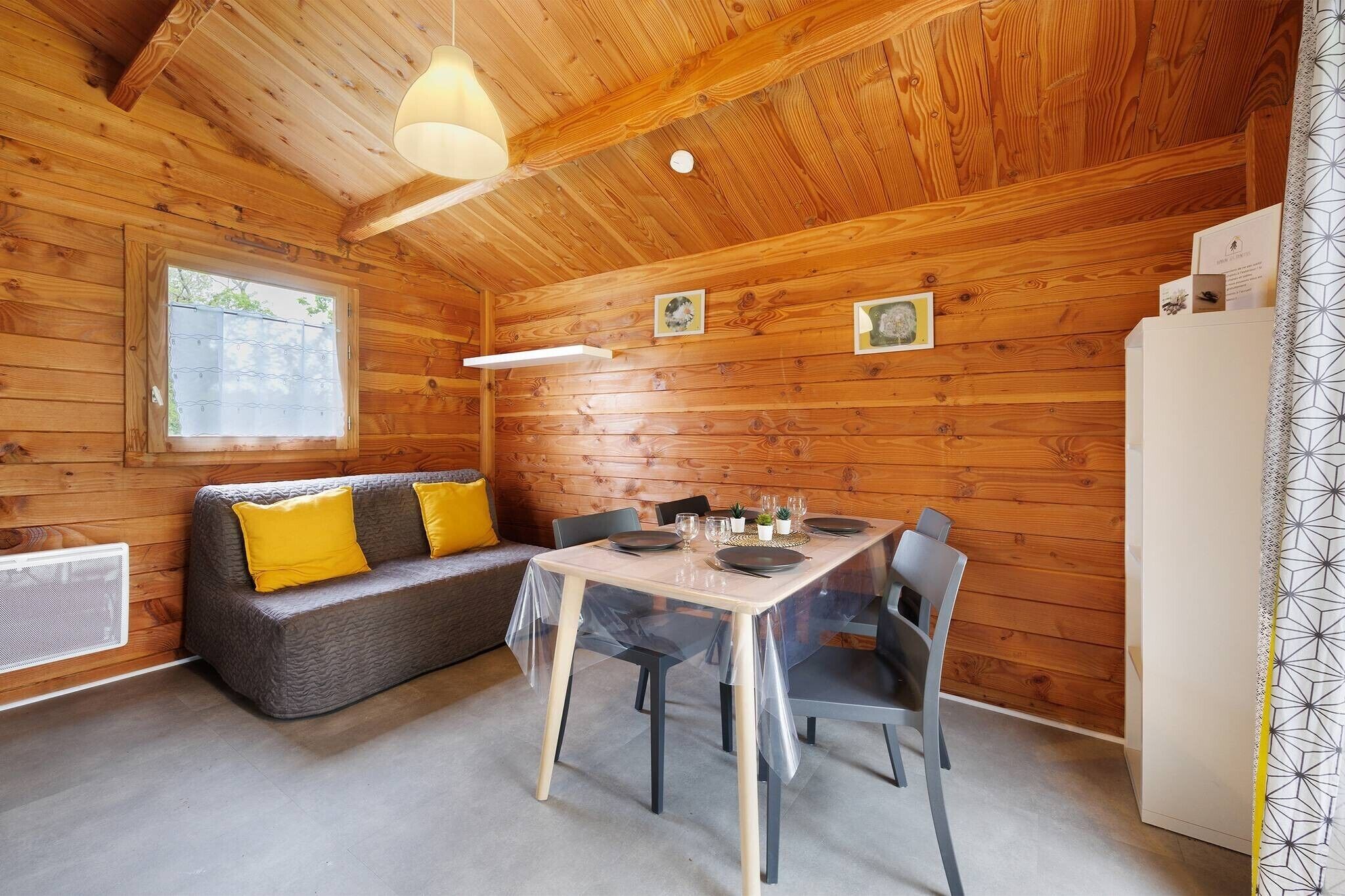 A wooden cabin interior with a dining table, chairs, a sofa with yellow cushions, a window, and a refrigerator.