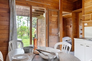 A wooden cabin interior with a dining table set for two, a kitchen area, and a sliding glass door leading to a garden. - Chalet “Bois 9” with Shared Pool and Private Terrace (Saint-Gervais)