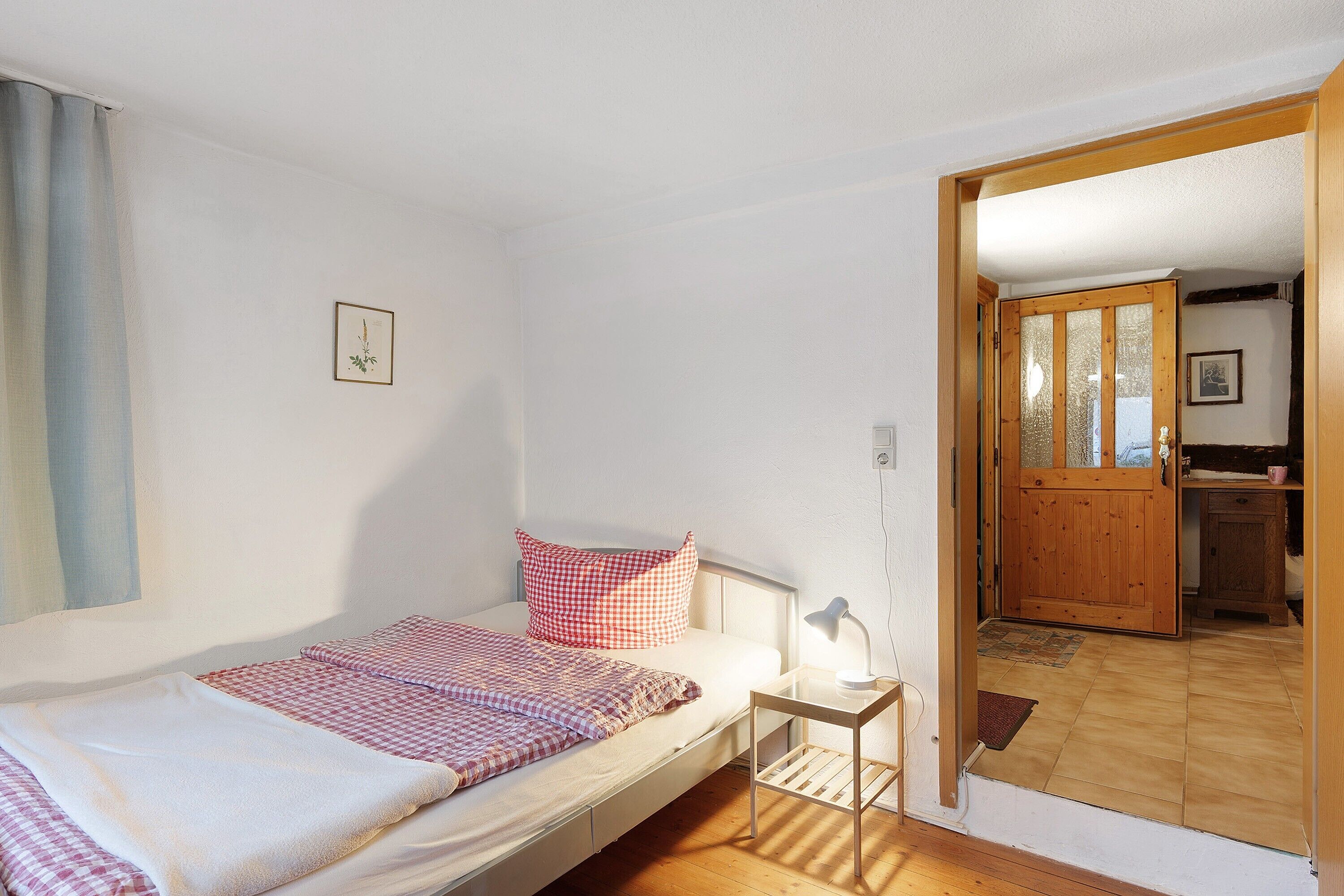Ein Einzelbett mit karierter Decke, ein Nachttisch mit Lampe und eine Holztür zu einem Badezimmer.