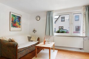 A living room with a sofa, a small table with a bottle, a clock, and a window with curtains. - Apartment 'Ferienwohnung 2' with Private Garden and Wi-Fi (Sternenfels)