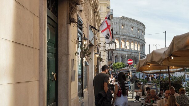 A street with a cafe, outdoor seating, and a historic building in the background. - DEZA Colosseum view (Roma)