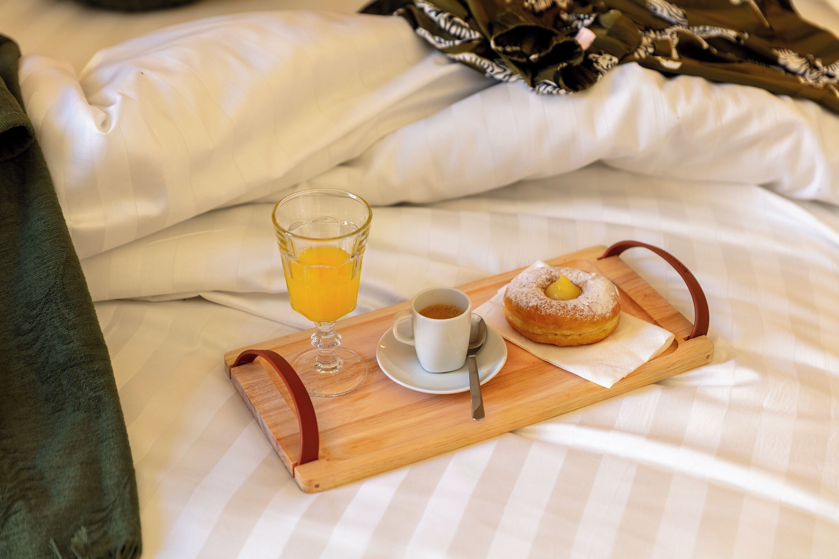 A breakfast tray with a glass of orange juice, a cup of coffee, and a donut on a bed with white bedding.