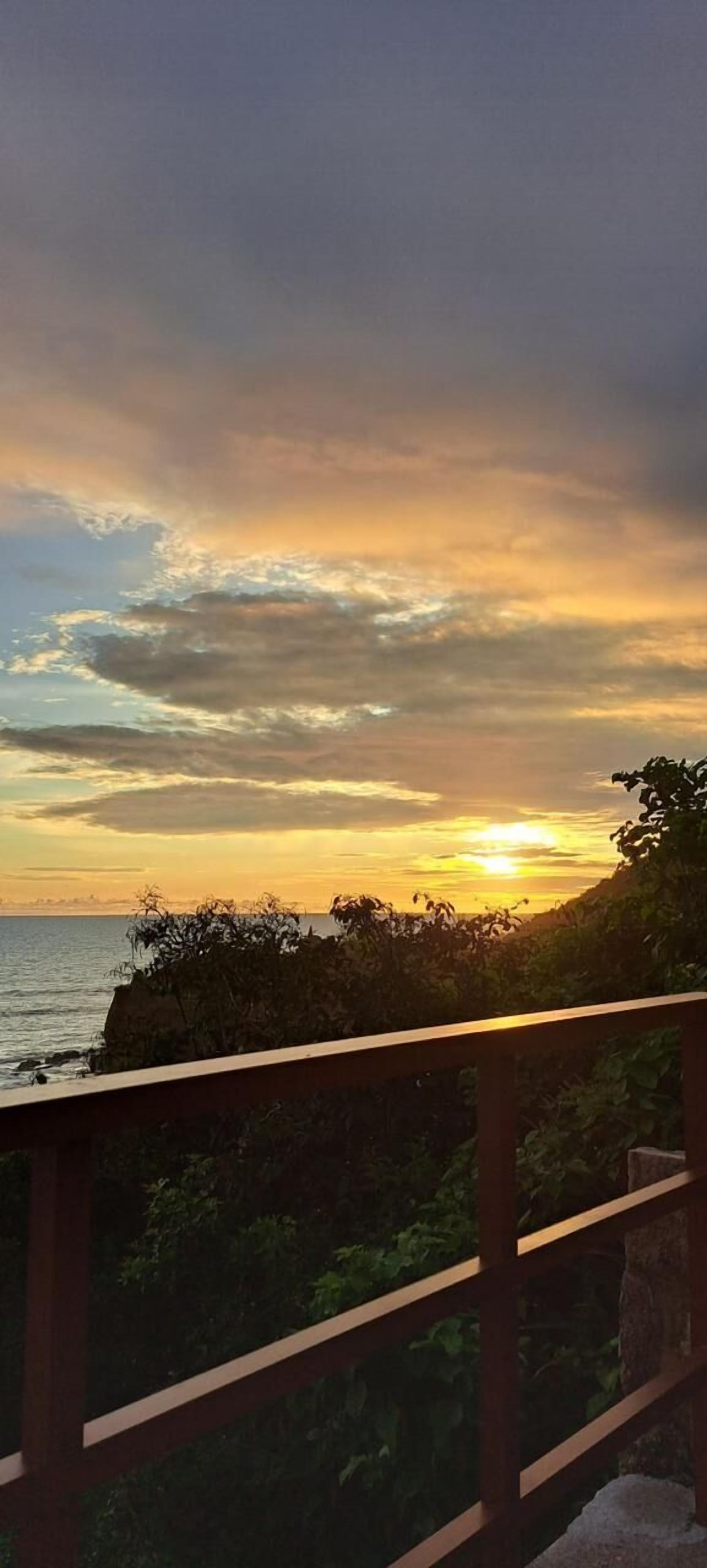 Vista del atardecer desde un balcón con barandilla, con vista a una masa de agua y una costa lejana.