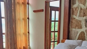 Una cama bien hecha con una manta color beige y ropa blanca, una pared de piedra y una ventana con cortinas.