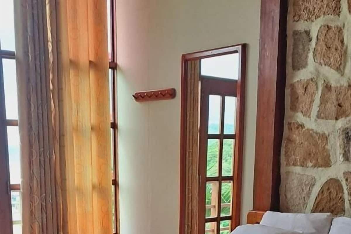 A neatly made bed with a beige blanket and white linens, a stone wall, and a window with curtains.