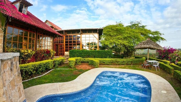 Una piscina con borde de piedra, rodeada por un jardín cuidado y una casa con ventanas grandes y tejado rojo.