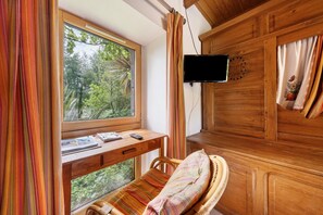 A room with a wooden desk, a chair, a bed with a striped blanket, a TV mounted on the wall, and a window with curtains.