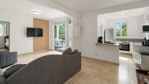 A modern, open-plan living space with a grey sofa, a flat-screen TV, a kitchen area, and a balcony with a view of greenery. - Villa Blanc de blanc (Les Anses-d'Arlet)