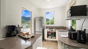A modern kitchen with a refrigerator, microwave, and coffee maker. There are windows providing a view of the mountains. - Villa Blanc de blanc (Les Anses-d'Arlet)
