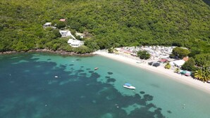 An aerial view of a coastal area with a beach, clear turquoise waters, and a small boat. There are buildings on the hillside and a cluster of structures near the shore. - Villa Blanc de blanc (Les Anses-d'Arlet)