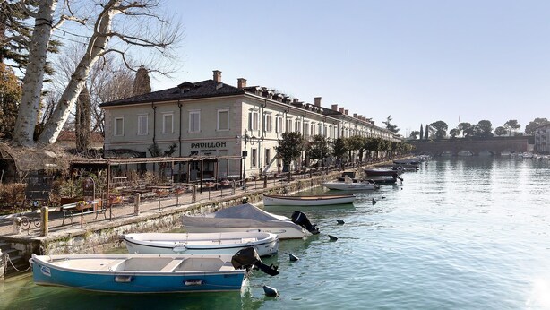 Une rangée de bateaux est amarrée le long d’une promenade au bord de l’eau, un bâtiment affiche une enseigne sur laquelle on peut lire « PAVILLON », et le ciel est dégagé.