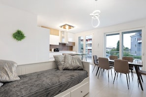 Ein modernes Schlafzimmer mit Bett, Essbereich mit Stühlen, Küchenzeile und Blick durch große Fenster.