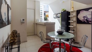Un espace repas moderne avec une table en verre, des chaises en métal et un tapis rouge. On aperçoit à l’arrière-plan un coin cuisine avec un comptoir et des étagères.