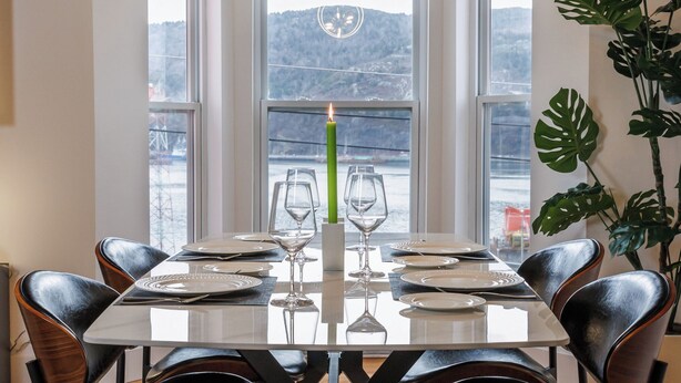 Ein Essbereich mit einem großen Tisch, gedeckt für sechs Personen, umgeben von Ledersesseln, mit Blick auf eine gebirgige Landschaft durch große Fenster.