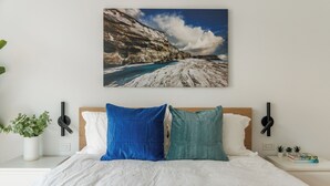 Ein Schlafzimmer mit einem Bett, Kissen, einem Nachttisch mit Pflanze, einer Wandlampe und einem Bilderrahmen mit einer Küstenlandschaft.
