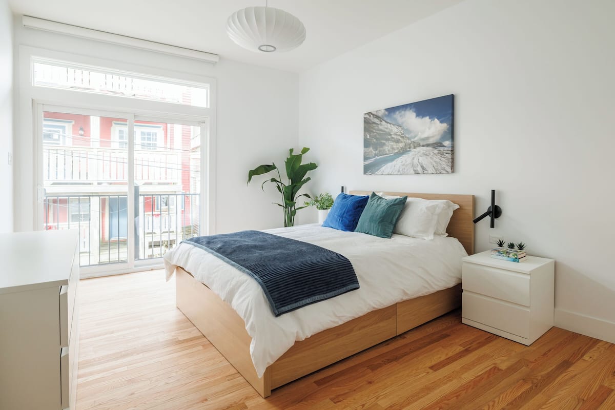 A bedroom with a bed, bedside table, and a window with a view of buildings.