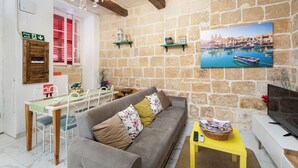 A cozy living room with a stone wall, a sofa, a dining table, and a television. - Traditional Townhouse - Rooftop Seaviews (Cospicua)