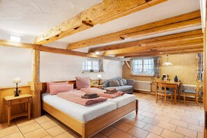 Um quarto com teto de madeira, cama com roupa de cama xadrez, mesa de cabeceira com abajur, janela com cortinas e uma pequena mesa com uma cadeira.