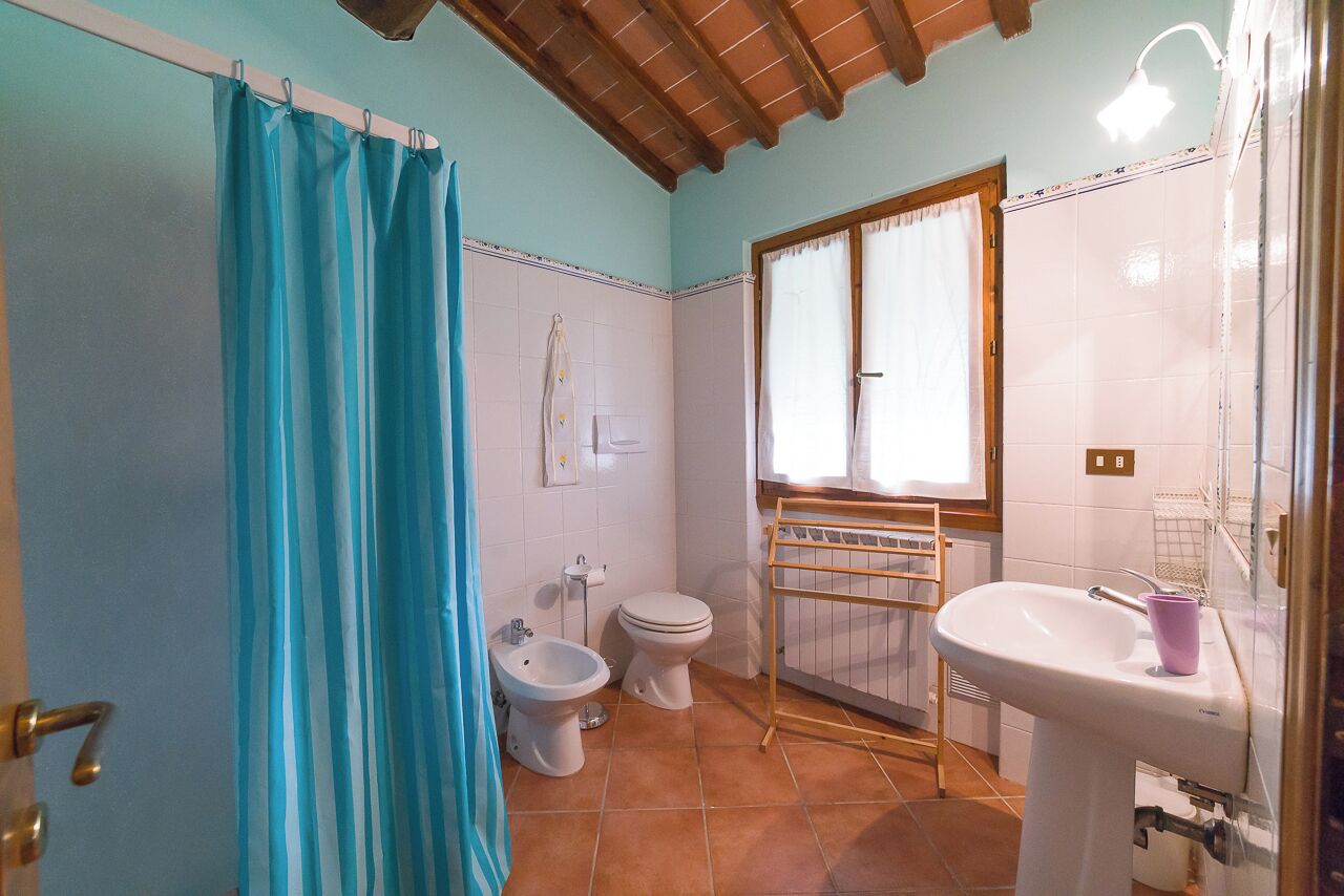A bathroom with a shower curtain, a bidet, a toilet, a window with curtains, and a sink with a mirror.