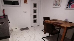 Een keuken met een koelkast, een eettafel met stoelen, een witte deur met een raam en een klein raam boven de deur.