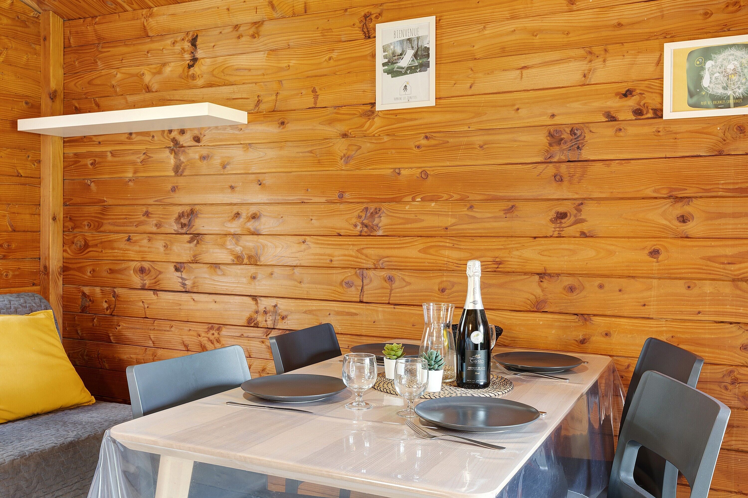 A wooden cabin interior with a dining table set for four, a bottle of champagne, and a framed welcome sign.