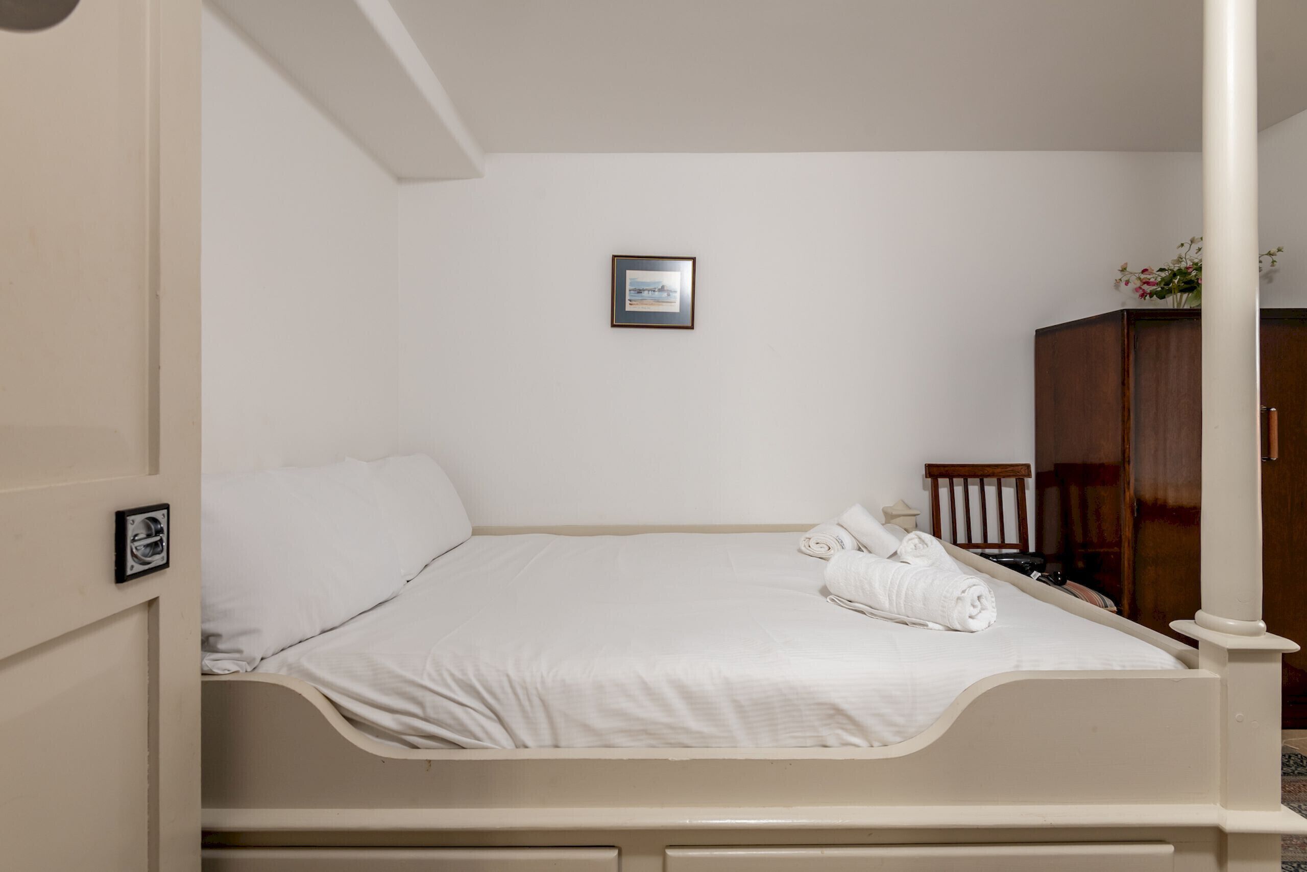 A neatly made bed with white linens and a rolled towel, a framed picture on the wall, and a wooden wardrobe.