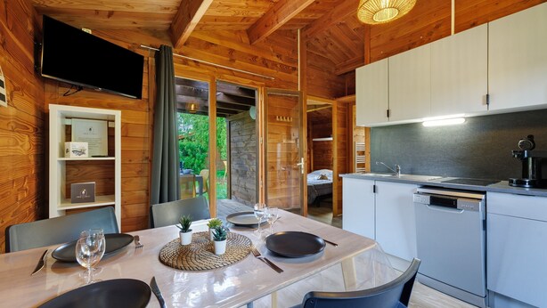 Een houten chalet van binnen, met een gedekte eettafel, een keuken en een aan de muur bevestigde televisie.