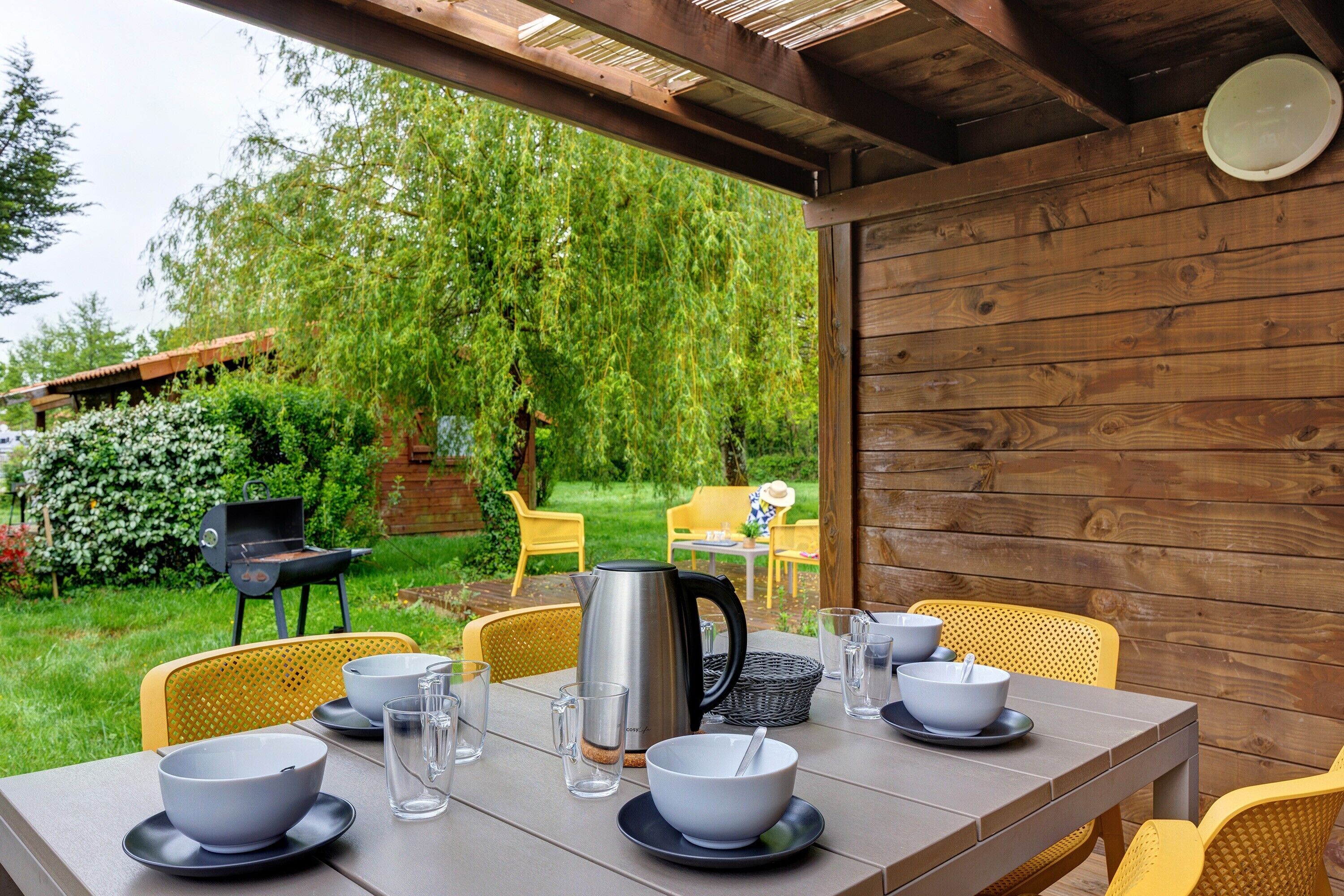 Een buiten eethoek met een tafel voor vier personen, omringd door gele stoelen, een metalen waterkoker en glazen.
