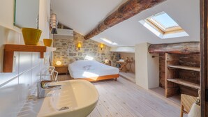 Un dormitorio con tragaluz, vigas de madera, pared de piedra y cama con colchón blanco.