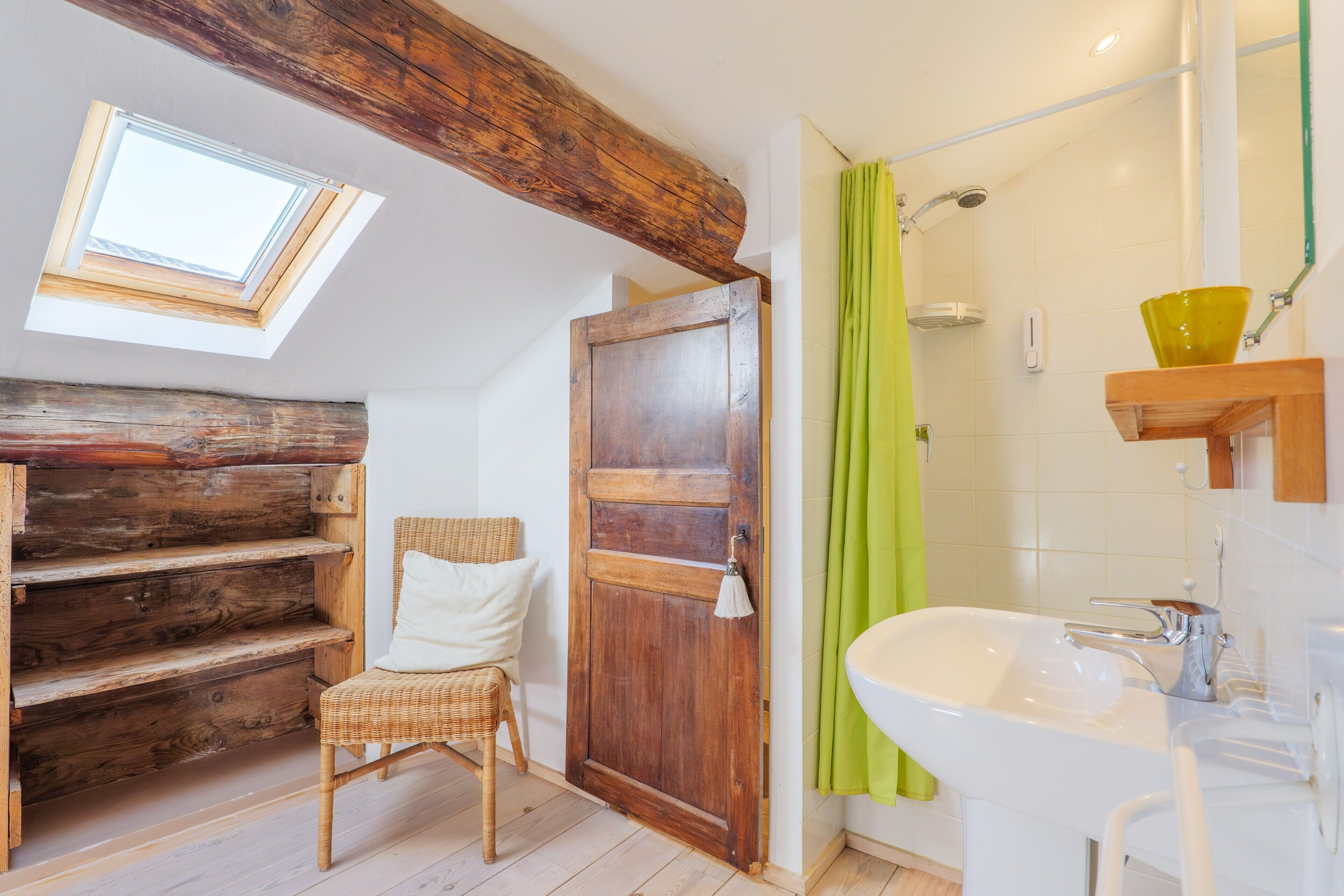 Ein Badezimmer mit Oberlicht, Holzbalken, einem Korbsessel, einer Holztür, einer Dusche mit grüner Vorhang und einem weißen Waschbecken.