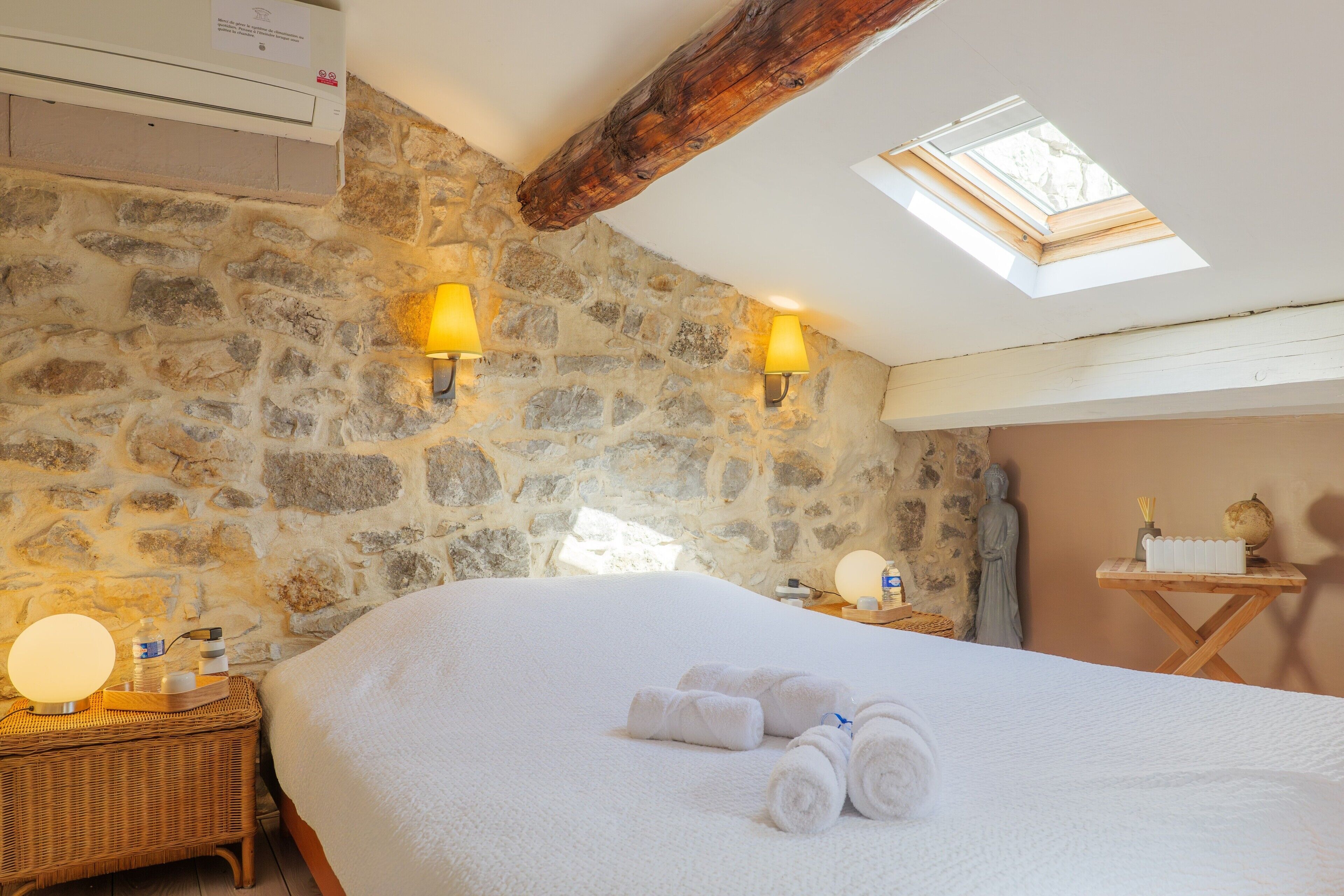 A room with a stone wall, a bed with white linens, two wall-mounted lamps, a wooden ceiling beam, and a skylight.