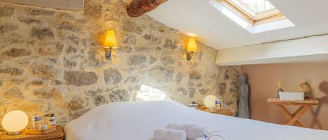 A room with a stone wall, a bed with white linens, two wall-mounted lamps, a wooden ceiling beam, and a skylight.