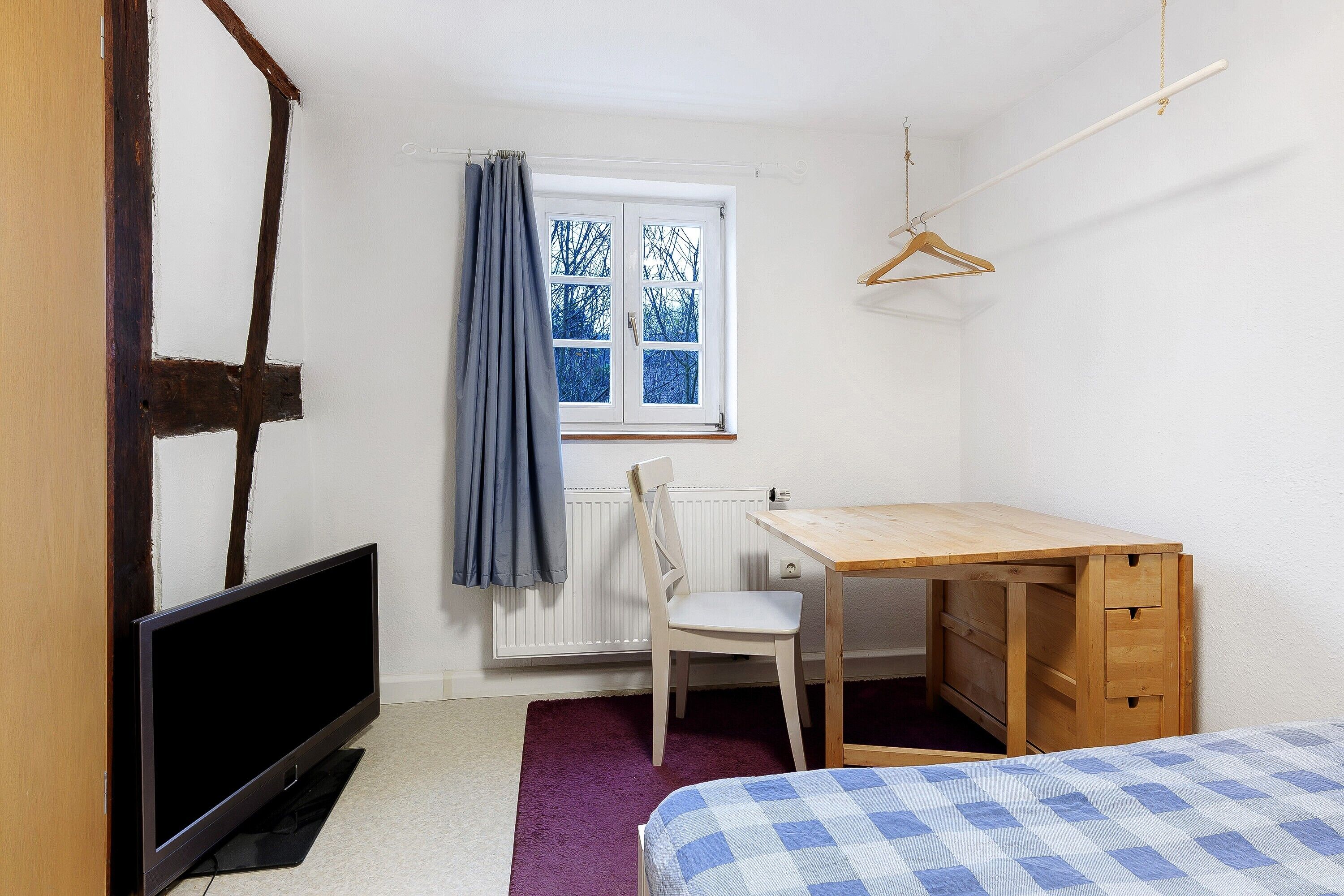 A room with a bed, a desk, a chair, a TV, and a window with a curtain.