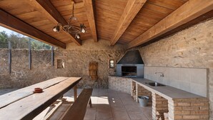A rustic outdoor kitchen with a stone wall, wooden beams, and a built-in grill. - Casa Rural La Moixella: Catalan Country House with Private Pool and Pyrenees Views (Lladurs)