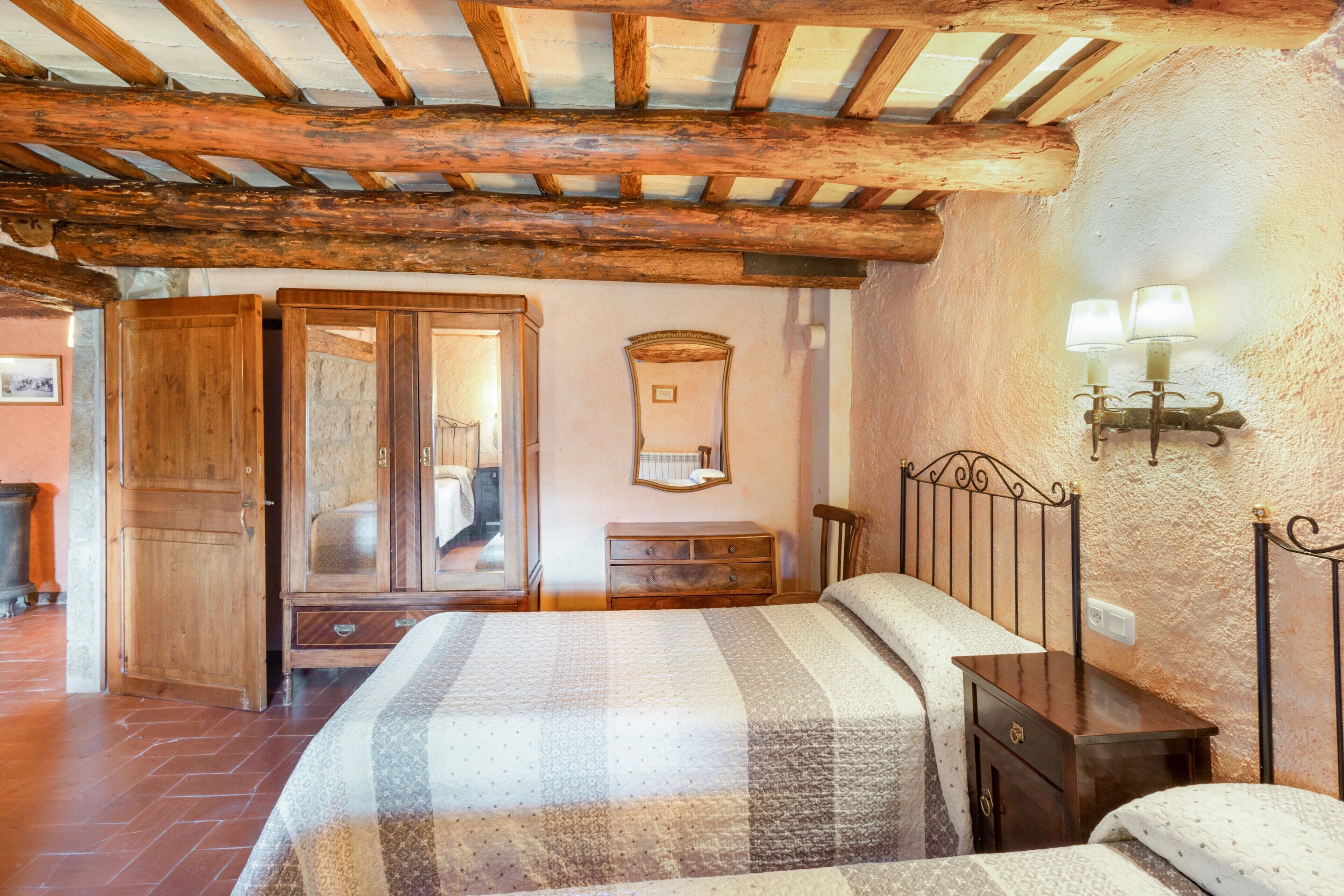 A bedroom with a wooden ceiling, a bed with a patterned bedspread, a dresser, a nightstand, and a wall-mounted lamp.