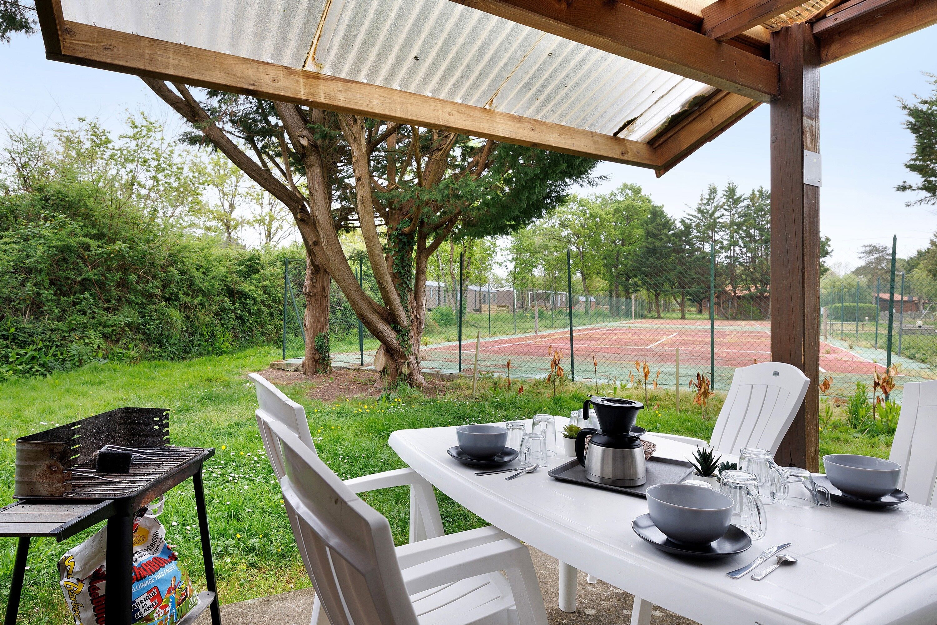 An outdoor dining area with a table set for two, surrounded by greenery and a tennis court in the background.