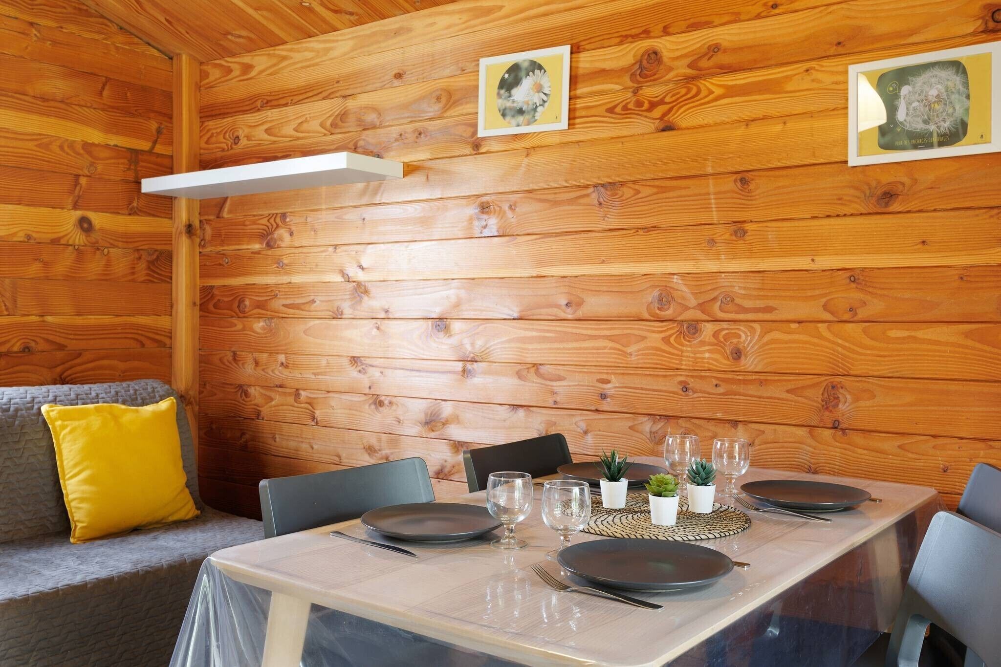 A wooden interior with a dining table set for four, chairs, a sofa with a yellow pillow, and framed pictures on the wall.