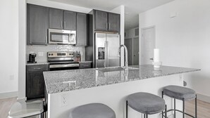 A modern kitchen with dark cabinets, stainless steel appliances, a granite countertop, and bar stools. - Landing | Amazing 2BD, Gym, Pool (Austin)