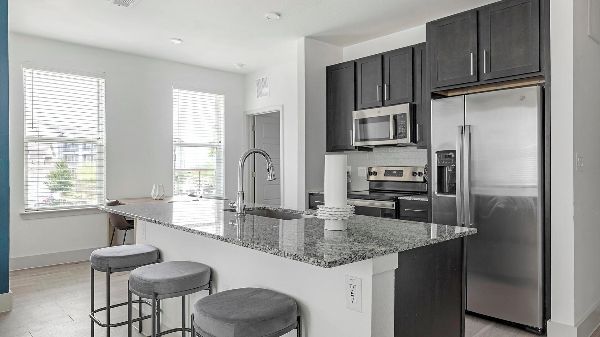 A modern kitchen with dark cabinets, stainless steel appliances, a granite countertop, and bar stools.