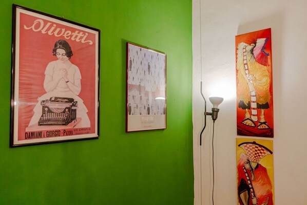 A green wall with three framed items: a vintage Olivetti advertisement, a framed abstract art piece, and a framed artwork with a checkered pattern.