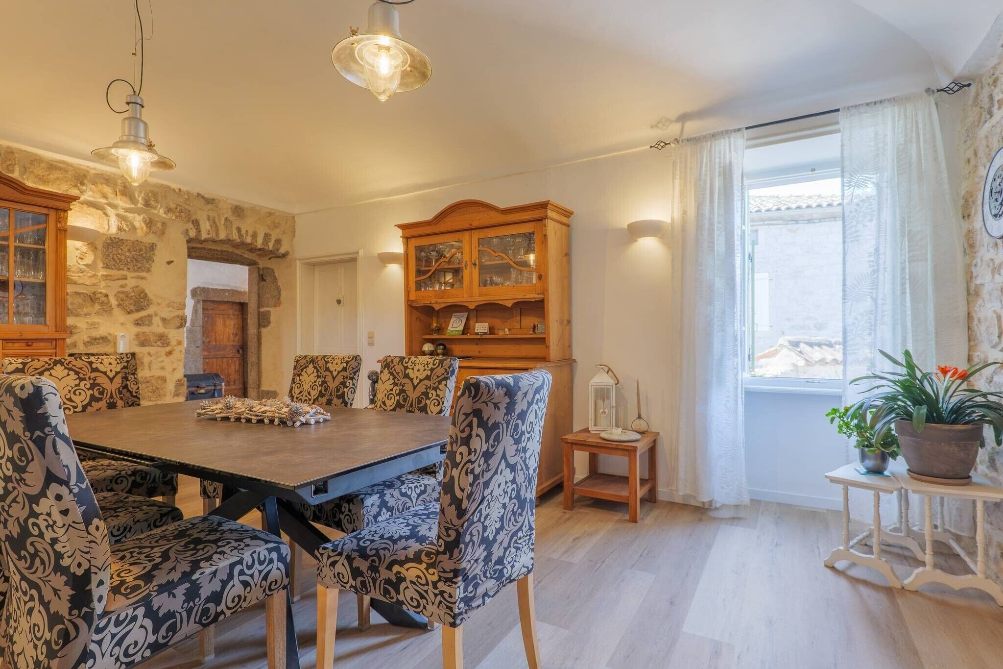 Un coin repas avec une table en bois et des chaises à motifs, un mur en pierre, un buffet en bois avec des portes en verre et une fenêtre avec des rideaux.