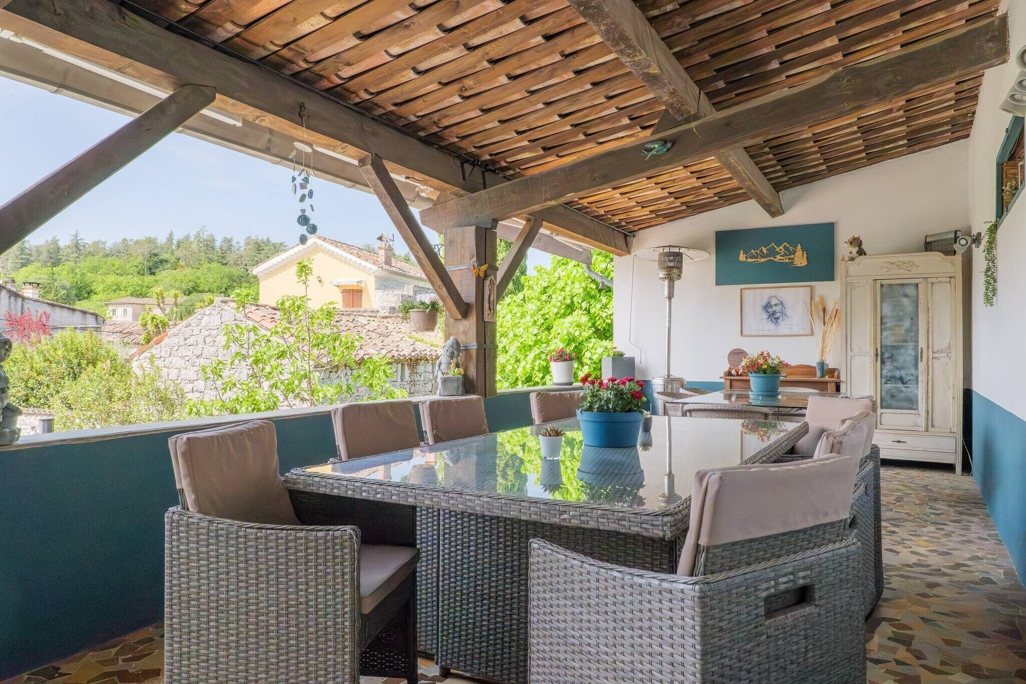 An outdoor patio with a glass-top table surrounded by wicker chairs, a wooden ceiling, and a view of the surrounding landscape.