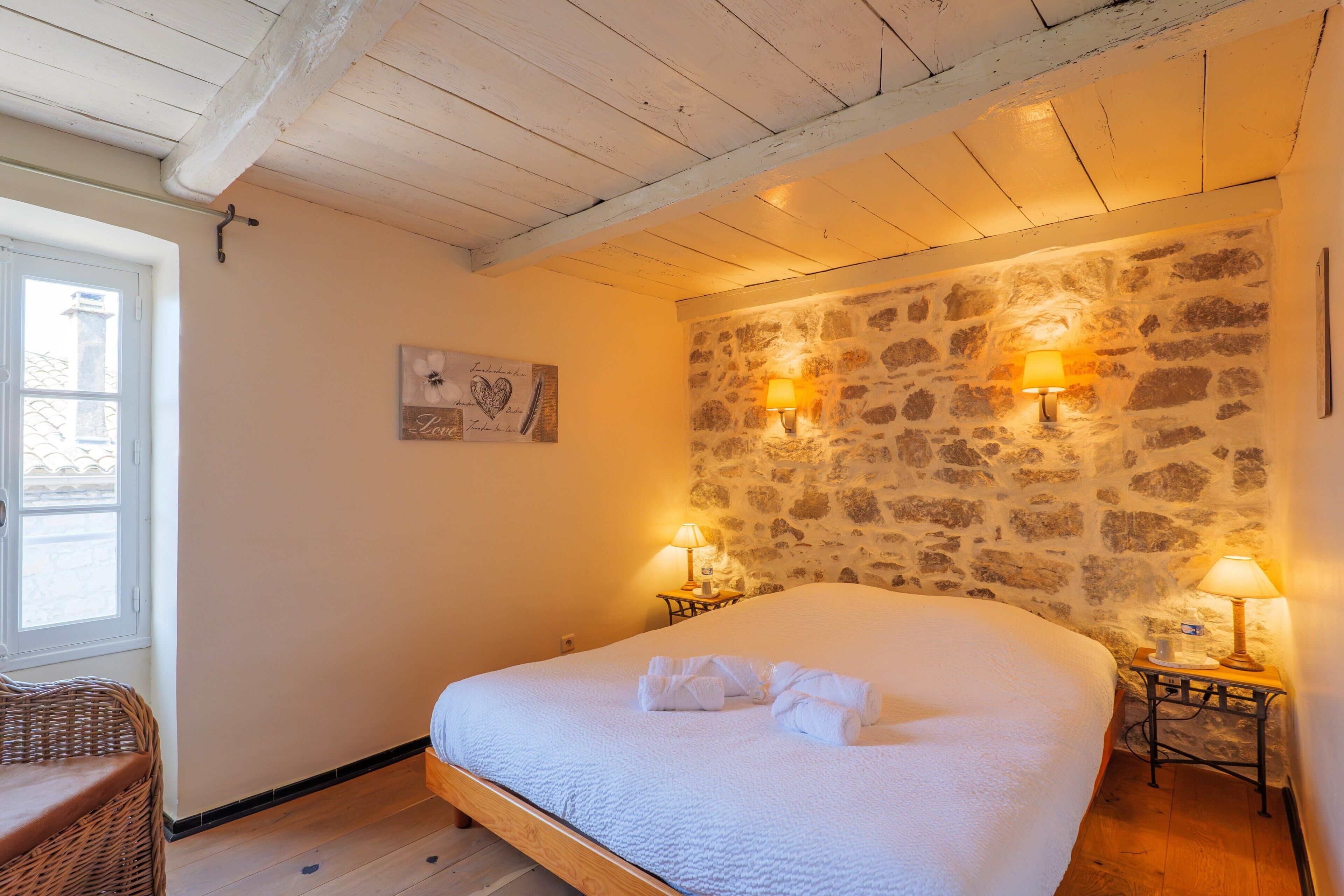 Ein Schlafzimmer mit Steinsturz, zwei Nachttischlampen, einem Bett mit weißer Bettwäsche und einem Korbstuhl.