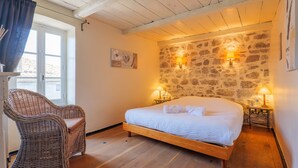 Un dormitorio con cama de madera, una silla de mimbre, pared de piedra y dos lámparas fijadas a la pared.