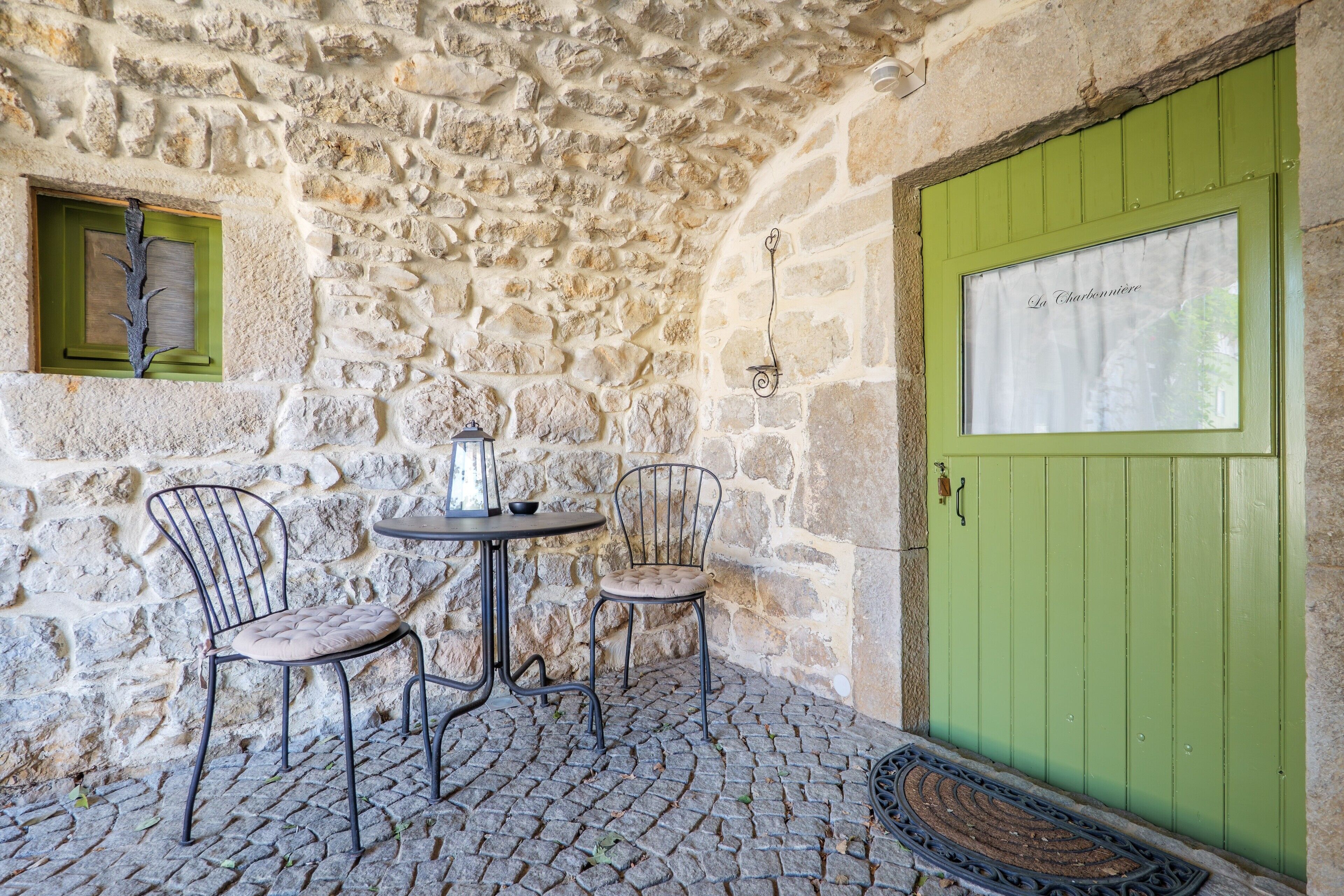 Un intérieur avec un mur en pierre, une porte verte, deux chaises en métal, une petite table et une fenêtre avec un cadre vert.