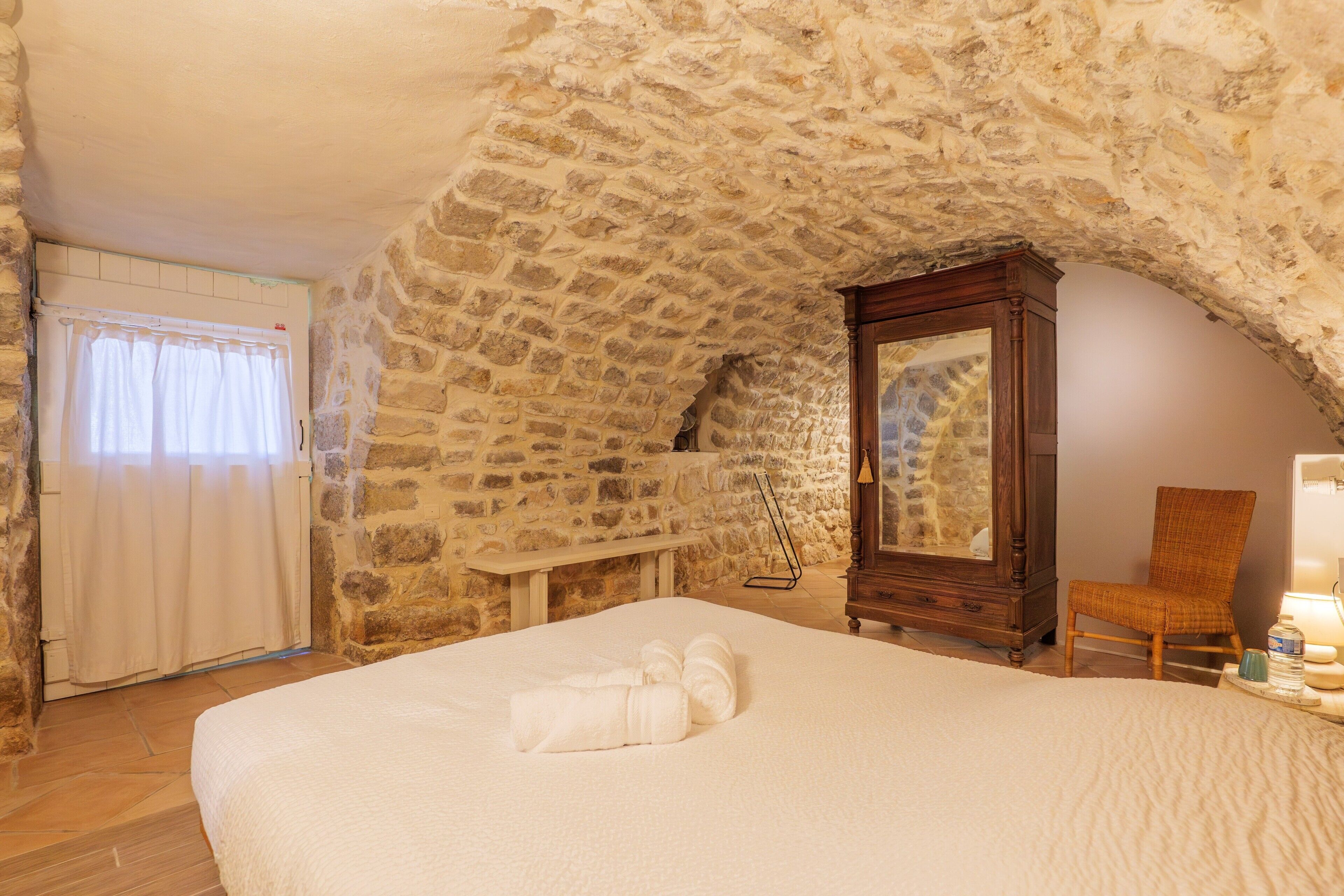 A bedroom with a stone ceiling, a bed with white linens, a wooden dresser, a chair, and a bedside table.