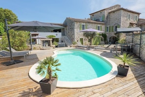 A swimming pool with a wooden deck, stone buildings, and outdoor furniture.