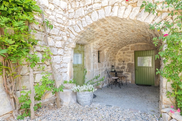 An arched stone structure with a table and chairs, a door, and potted plants.