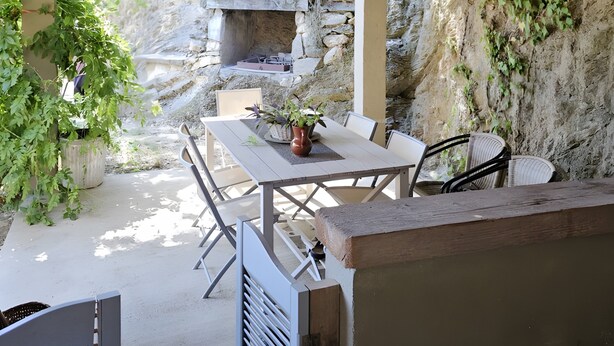 Um pátio externo com mesa e cadeiras, um candeeiro pendente e uma parede de pedra com vegetação.