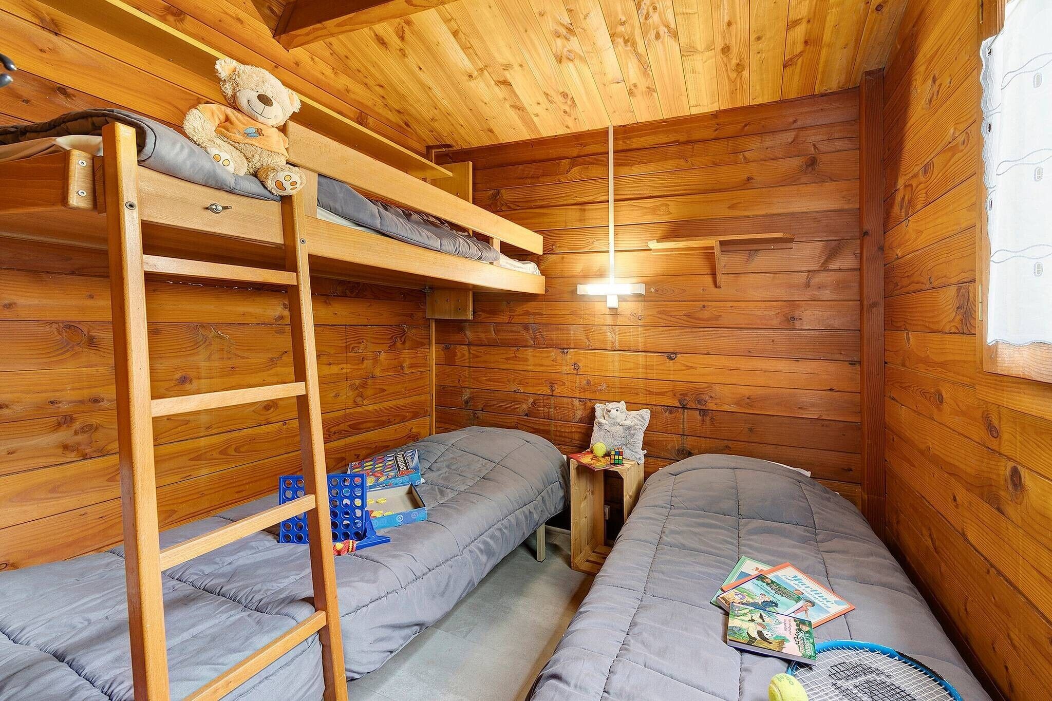 A wooden cabin room with bunk beds, a ladder, a small table with a teddy bear, and a window with curtains.